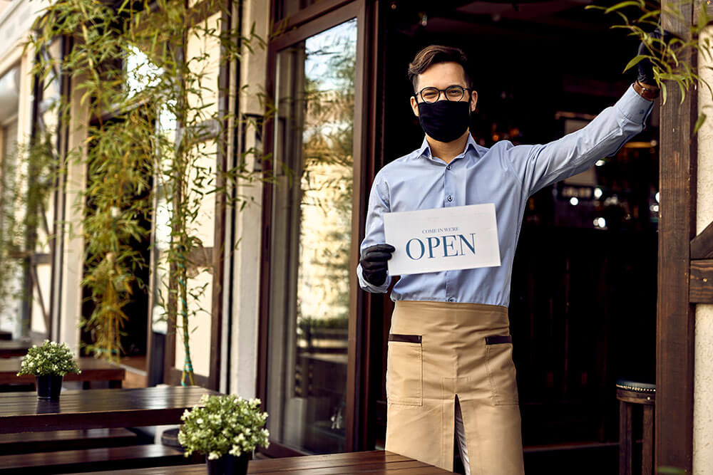 capitalist financial man in an apron standing in front of a restaurant entrance smiling while holding a sign that says come in we're open